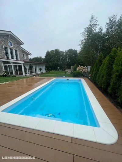 Прямоугольный бассейн с голубой водой и террасой из коричневого материала у каменного дома